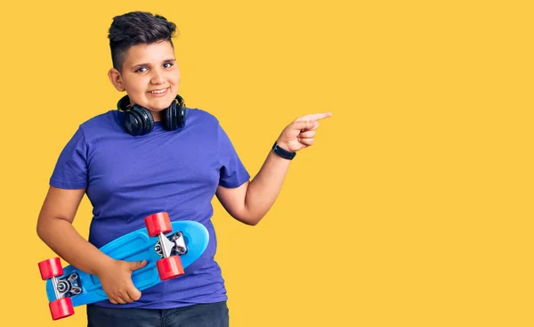 小さな男の子保持スケートと身に着けているヘッドフォン笑顔幸せなポインティングで手と指で側面 — ストック写真