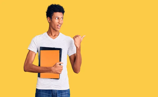 Joven Afroamericano Hombre Sosteniendo Libro Apuntando Pulgar Hacia Lado Sonriendo —  Fotos de Stock