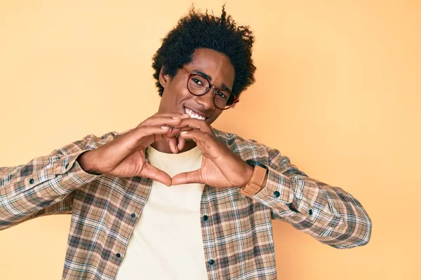 Handsome african american man with afro hair wearing casual clothes and glasses smiling in love doing heart symbol shape with hands. romantic concept.