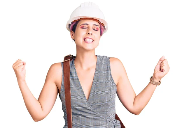 年轻美丽的女人戴着建筑师的硬礼帽 头戴皮包 非常快乐而兴奋地举起双臂做着获胜的手势 微笑着 尖叫着要成功 庆祝概念 — 图库照片
