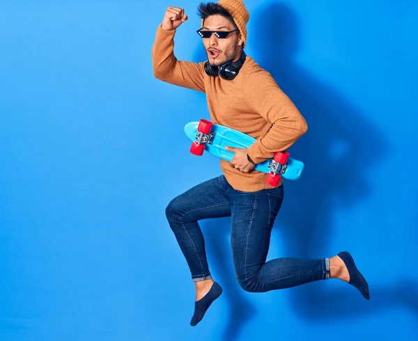 Young handsome latin man wearing wool cap and thug life glasses.  Jumping with smile on face holding skate over isolated blue background