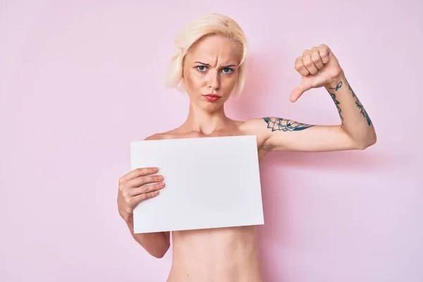 Young blonde woman with tattoo standing shirtless holding empty blank banner with angry face, negative sign showing dislike with thumbs down, rejection concept