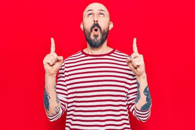 Gündelik giysiler giyen genç yakışıklı adam şaşırmış ve şaşırmış bir şekilde yukarı bakıyor ve parmaklarıyla işaret ediyor ve kollarını kaldırıyor.. 