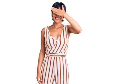 Kısa saçlı, kısa saçlı, yaz elbisesi giyen, gülümseyen ve elleri yüzünde, şaşırtmak için gözlerini kapatan genç esmer kadın. kör kavram. 