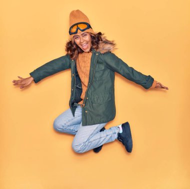 Kışlık giysiler giyen genç, güzel Latin kadın mutlu gülümsüyor. Yüzünde ve kollarında tebessümle, izole edilmiş sarı arka planda atlıyor.