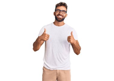 Gündelik kıyafetler giyen ve gözlük takan genç İspanyol adam. Başarıyla olumlu jest yapıyor. Gülümsüyor ve mutlu. Neşeli ifade ve kazanan jest. 