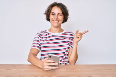 Genç İspanyol kadın akıllı telefon kullanıyor masada oturuyor gülümsüyor mutlu bir şekilde el ve parmak kenarı işaret ediyor. 