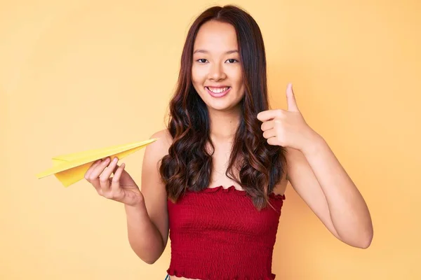 年轻美丽的中国姑娘牵着纸飞机 笑容满面 大拇指向上做得很出色 签了字 — 图库照片