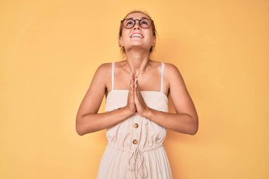 Yazlık elbiseler giyen beyaz güzel bir kadın elleri bağlı yalvarıyor ve dua ediyor. Yüzünde umut ifadesi var. Çok duygusal ve endişeli. Yalvarıyor. 