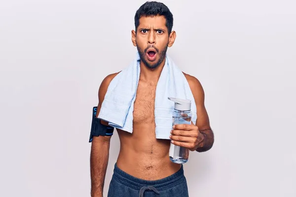 年轻的拉丁男子身穿运动服 手拿着毛巾 手里拿着一瓶水 惊慌失措 张大了嘴 满脸诧异 — 图库照片