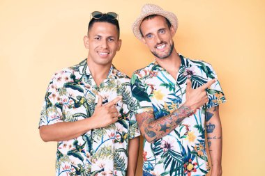 Yazlık şapkalı ve Hawaii tişörtlü iki genç eşcinsel. Yüzünde bir gülümseme, mutlu ve doğal bir ifadeyle elini ve parmağını kaldırmış. 