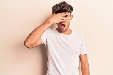 Gündelik kıyafetler giyen genç İspanyol adam şok içinde yüzünü ve gözlerini eliyle kapatıyor, parmaklarından korkuyla bakıyor. 