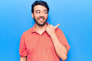 Gündelik kıyafetler giyen, gülümseyen, gülümseyen ve başparmağıyla yan tarafı gösteren İspanyol genç adam.. 