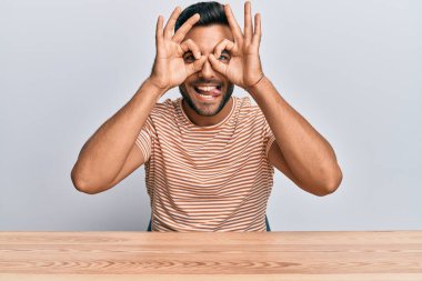 Masada oturan, sıradan kıyafetler giyen yakışıklı İspanyol bir adam. Dürbün gibi jest yapıyor, gözleri parmaklarından bakıyor. çılgın ifade. 