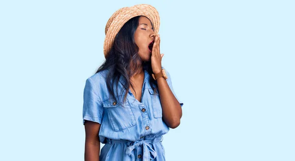 Young indian girl wearing summer hat bored yawning tired covering mouth with hand. restless and sleepiness.