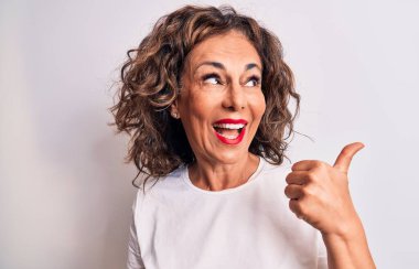 Orta yaşlı, güzel esmer kadın, günlük tişörtler giyip beyaz arka planda dikiliyor başparmağıyla yan tarafa bakıyor ve ağzı açık gülümsüyor.