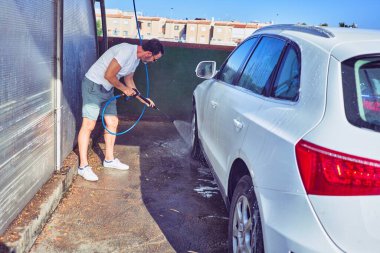 Orta yaşlı yakışıklı, günlük kıyafetler giyen ve mutlu gülümseyen güneş gözlüklü bir adam. Yüzünde gülümsemeyle ayakta durup su tabancası kullanarak araba yıkamak..