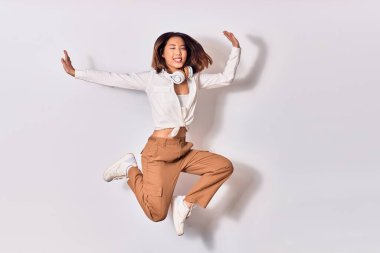 Genç, güzel Çinli kadın mutlu gülümsüyor. Yüzünde gülümsemeyle zıplıyor, beyaz arka planda kulaklık kullanarak müzik dinliyor.