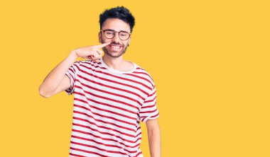 Latin asıllı genç bir adam, gündelik giysiler giyip el ve burnu işaret ediyor, neşeli bir şekilde gülümsüyor. güzellik konsepti 