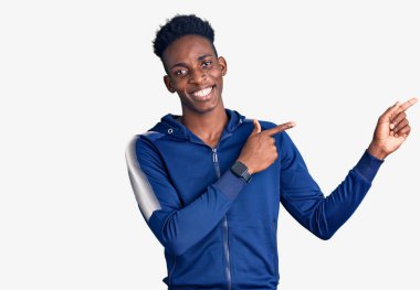 Afro-Amerikalı genç adam spor kıyafetleri giyiyor gülümsüyor ve kameraya bakıyor iki eliyle ve parmaklarıyla kenarı işaret ediyor.. 