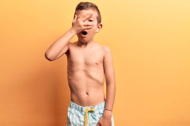 Mayo giyen sevimli sarışın çocuk şok içinde yüzünü ve gözlerini kapatıyor, parmaklarından korkuyla bakıyor. 