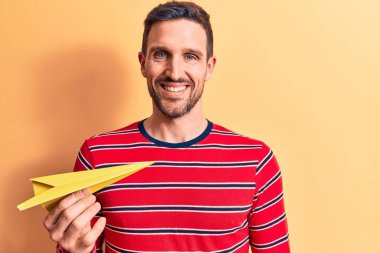 Sarı kağıt uçak tutan yakışıklı genç adam izole edilmiş arka planda duruyor ve pozitif ve mutlu görünüyor kendine güvenen bir gülümsemeyle dişlerini gösteriyor.