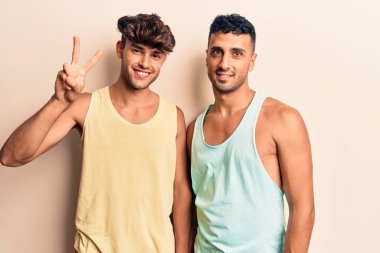 Genç eşcinsel çift gündelik giysiler giyip iki numarayı gösteriyorlar ve mutlu bir şekilde gülümsüyorlar.. 