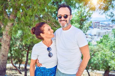 Orta yaşlı, güzel bir çift, günlük kıyafetler ve mutlu bir şekilde gülümseyen güneş gözlüğü takıyor. Yüzünde gülümsemeyle ayakta durup ormanda sarılıyor.