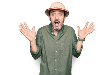 Tatilde olan orta yaşlı bir turist, izole edilmiş beyaz arka plan üzerinde kaşif şapkası takıyor ve kolları açık, hiçbir fikri yok ve şüpheli bir yüzü var..