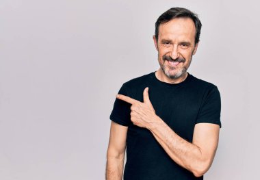 Sıradan siyah tişört giyen orta yaşlı bir adam izole edilmiş beyaz arka planda dikiliyor. Gülümsüyor, eli ve parmağı yana doğru uzanıyor.