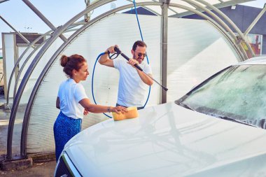 Orta yaşlı, güzel bir çift günlük kıyafetler giyip mutlu gülümsüyor. Yüzünde gülümseme, su tabancası ve sünger kullanarak araba yıkamak..