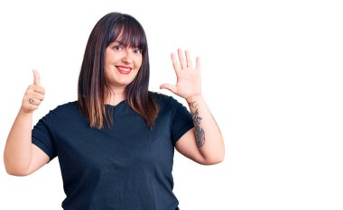 Genç ve cüsseli kadın gündelik giysiler giyiyor. Altıncı parmaklarını gösterip işaret ederken kendinden emin ve mutlu gülümsüyor.. 