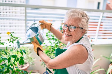 Gri saçlı, eldivenli ve bahçıvan önlüklü yaşlı kadın evdeki bitkileri bahçeliyor.