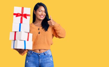 Uzun saçlı İspanyol kadın elinde hediyeler mutlu ve pozitif gülümsüyor, başparmağıyla mükemmel ve onay işareti yapıyor. 