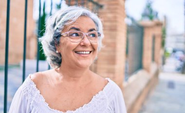 Beyaz saçlı orta yaşlı bir kadın mutlu bir şekilde dışarıda gülümsüyor.