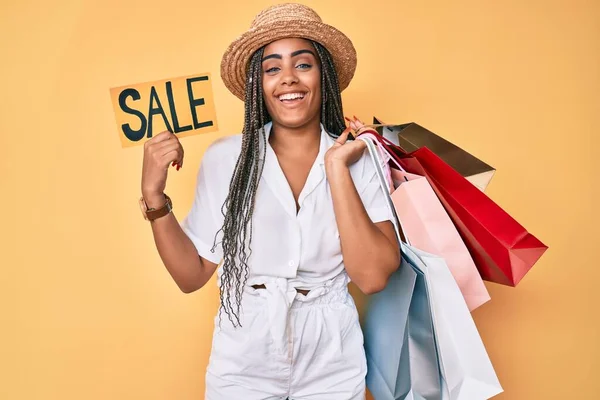 Young african american woman with braids holding shopping bags and sale banner smiling and laughing hard out loud because funny crazy joke.