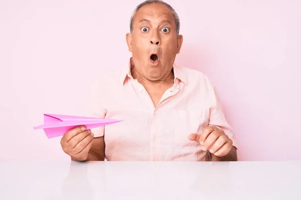 Senior handsome man with gray hair holding paper airplane scared and amazed with open mouth for surprise, disbelief face