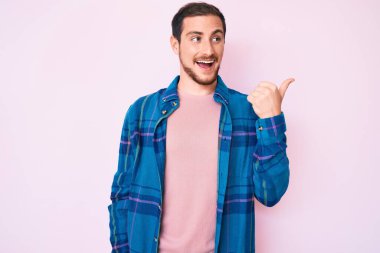 Genç, yakışıklı, sıradan kıyafetler giyen, gülen yüzlü, gülümseyen ve baş parmağıyla yan tarafı gösteren bir adam.. 