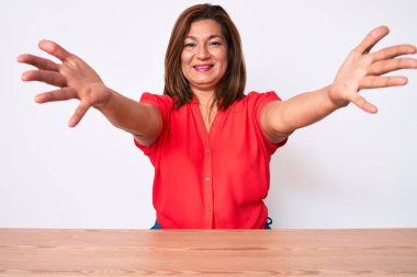 Orta yaşlı esmer kadın, gündelik giysiler giymiş masaya oturmuş kameraya bakıyor ve sarılmak için kollarını açmış gülümsüyor. Mutluluğu kucaklayan neşeli bir ifade.. 