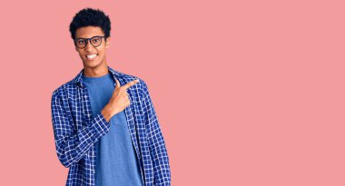 Genç Afro-Amerikan adam, günlük kıyafetler ve gözlük takıyor. Neşeli, yüzünde mutlu ve doğal bir ifadeyle, elleri ve parmaklarıyla yüzünü gösteren bir gülümseme var. 