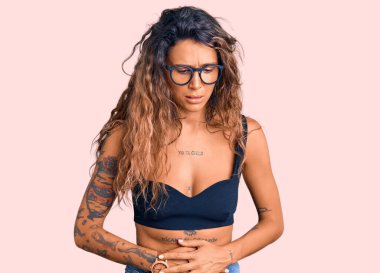 Dövmeli genç İspanyol kadın günlük kıyafetler giyiyor ve elleri karnında gözlükler takıyor çünkü hazımsızlık, acı verici bir hastalık kendini iyi hissetmiyor. Ağrı kavramı. 