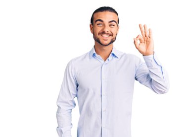 İş kıyafetleri giymiş yakışıklı bir genç pozitif gülümsüyor ve el ve parmaklarıyla imza atıyor. başarılı ifade. 