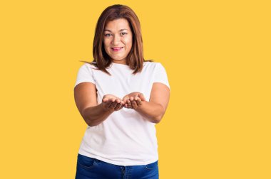 Orta yaşlı, beyaz tişört giyen, elleri avuçlarında gülümseyen, el hareketi yapan Latin bir kadın. Bekleme ve koruma 