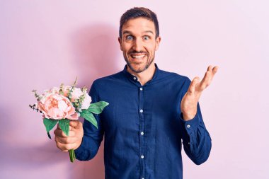 Genç yakışıklı romantik adam pembe arka planda güzel bir buket çiçek tutarak başarıyı mutlu bir gülümsemeyle ve kazanan yüz ifadesiyle el kaldırarak kutluyor.