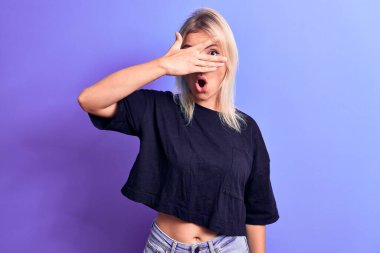 Genç, güzel, sarışın bir kadın, sıradan siyah bir tişört giyiyor, mor arka planda dikiliyor, şok içinde yüzünü ve gözlerini eliyle kapatıyor, parmaklarından korkuyla bakıyor.