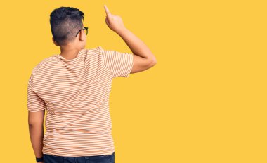 Gündelik giysiler ve gözlük takan küçük bir çocuk parmak eliyle öne doğru poz veriyor. 