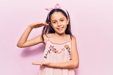 Güzel bir kız çocuğu günlük kıyafetler giyiyor. Elleriyle işaret ediyor. Büyük ve büyük beden işareti, ölçü işareti. Kameraya bakıp gülümsüyor. ölçüm kavramı. 