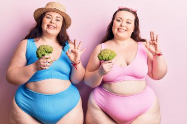 Bikini giyen, brokoli tutan, parmaklarıyla imza atan, gülümseyen, arkadaşça hareketler yapan, harika bir sembol olan genç ve büyük beden ikizler. 