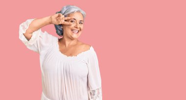 Gri saçlı, bohem tarzı giyinen, barış sembolü takan, yüzünde parmakları olan, gülümseyen, zaferi gösteren son sınıf öğrencisi bir kadın. 