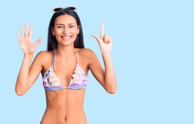 Bikini giyen, güneş gözlüklü genç ve güzel bir Latin kız yedi numaralı parmaklarını gösterip işaret ederken kendinden emin ve mutlu bir şekilde gülümsüyor.. 